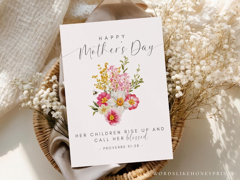 May include: A Mother's Day card with a floral bouquet design. The card features the text "Happy Mother's Day" and a quote from Proverbs 31:28. The bouquet includes pink, yellow, and white flowers with green foliage. The card is set on a woven basket.
