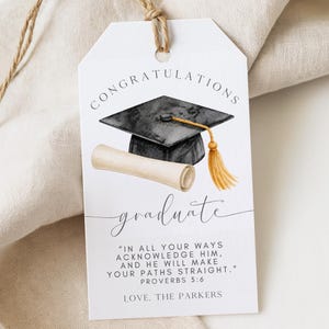 May include: A white graduation gift tag with the words "CONGRATULATIONS graduate" in elegant script. The tag features a watercolor illustration of a graduation cap, a diploma, and a tassel. The tag also includes a Bible verse.