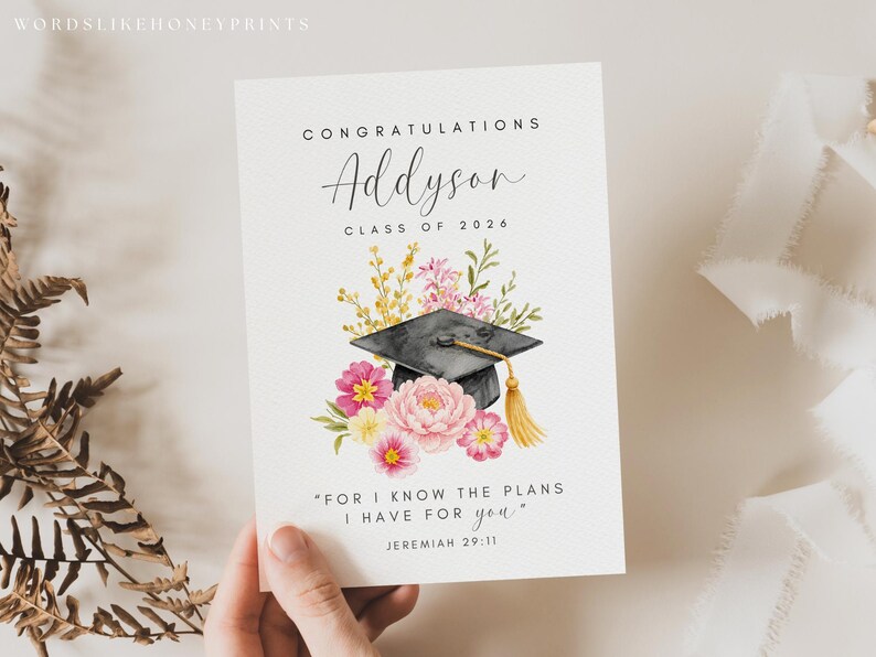 Puede incluir: Tarjeta de graduaci&oacute;n con dise&ntilde;o floral y birrete en acuarela. La tarjeta dice "Felicidades Addyson, Clase de 2026" e incluye una cita de Jerem&iacute;as 29:11. Flores rosas y amarillas rodean el birrete.
