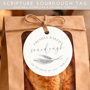 Könnte beinhalten: Eine braune Papiertüte mit einem transparenten Fenster, die ein Sauerteigbrot enthält. Ein rundes Etikett mit den Worten "FRESHLY BAKED sourdough" und "I AM THE BREAD OF LIFE." JOHN 6:35" ist mit Bindfaden an der Tüte befestigt.