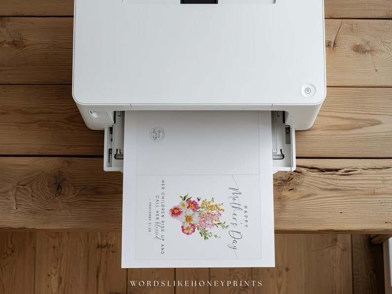 May include: A white printer printing a Mother's Day card. The card features a floral arrangement with pink, yellow, and white flowers, along with the text "Happy Mother's Day". The card also includes the text "Her children rise up and call her blessed".