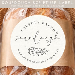 Può includere: Un'etichetta rotonda su una pagnotta di pane a lievitazione naturale. L'etichetta recita "FRESHLY BAKED sourdough" con un rametto di foglie e il testo "I AM THE BREAD OF LIFE." JOHN 6:35. L'etichetta è beige con testo nero.