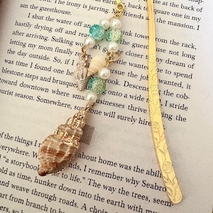 May include: A gold-colored metal bookmark with a seashell charm, pearls, and turquoise beads. The bookmark is on a book page with the text "JUST FRIENDS". The seashell charm is approximately 1 inch in length.