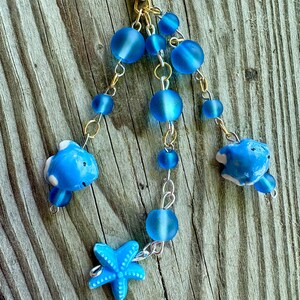 May include: A handmade keychain featuring blue beads, small blue fish charms, and a blue starfish charm. The keychain has gold and silver-colored chains and is set against a weathered wood background.