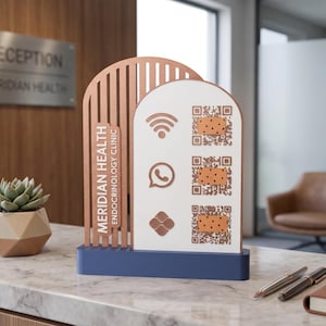 Pode incluir: Uma placa de mesa para Meridian Health Endocrinology Clinic com uma moldura arqueada cor de cobre com ripas verticais, um painel branco com três ícones e códigos QR, sobre uma base retangular azul-marinho sólida.