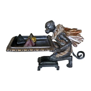 May include: A decorative monkey figurine kneeling on a small black base, holding a rectangular tray with a decorative border. The monkey has dark fur and a metallic cape. Three small pyramid-shaped objects in black, amber, and red sit on the tray.