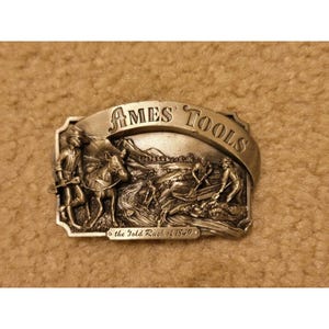 May include: A rectangular metal belt buckle with a silver-toned finish featuring a raised relief scene of prospectors and a pack animal. The text AMES TOOLS is arched across the top, and the text the Gold Rush of 1849 is centered at the bottom.
