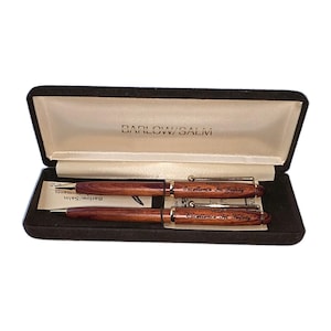 May include: A set of two dark wood pens with gold-tone clips and accents, presented in a black velvet-lined case. The pens are engraved with the text Excellence In Safety, and the interior of the case lid features the text BARLOW/SALM.