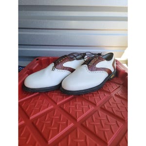 May include: A pair of white and brown leather saddle shoes featuring a textured crocodile-embossed pattern on the brown sections, dark brown laces, gold-tone eyelets, decorative brogue perforations, and a solid black rubber sole.