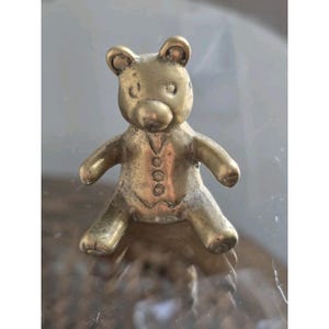 Könnte beinhalten: Eine kleine, goldfarbene Messingfigur eines sitzenden Teddybären. Der Bär hat einen glatten, abgerundeten Kopf mit kleinen Ohren und einen Rumpf, der mit einem eingravierten Westenmuster verziert ist, das drei runde Knöpfe auf der Vorderseite zeigt.