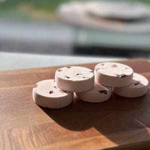 Peut inclure: Six bombes de bain rondes, rose pâle, avec des pétales de fleurs rouges visibles, sont empilées sur une planche en bois. Les bombes de bain sont disposées en quinconce, avec un arrière-plan flou.