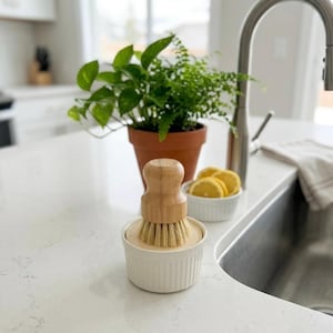 May include: A white ceramic dish brush with a wooden handle and natural bristles sits on a white countertop. A small bowl of lemon slices and a potted plant are in the background. The kitchen sink and tap are also visible.