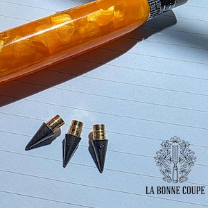 Puede incluir: Un portaminas de resina naranja y dorada con punta y goma plateadas, sobre un cuaderno rayado. Tres puntas de repuesto para lápiz están junto al lápiz. El lápiz tiene un aspecto veteado único. El logotipo "LA BONNE COUPE" está en la esquina inferior derecha.