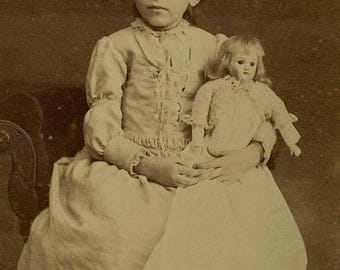 Antica fotografia su cartoncino vittoriano raffigurante una bambina che tiene in mano una bambola di porcellana (anni 1880).