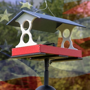 May include: A red, white, and blue bird feeder with a roof and a tray. The feeder is mounted on a black pole.