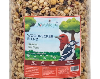 Może przedstawiać: Przezroczysty plastikowy cylinder wypełniony mieszanką nasion dla ptaków dla dzięciołów. Etykieta zawiera napis "JCs Wildlife Woodpecker Blend Premium Bird Seed" i ilustrację dzięcioła o czerwonej głowie. Etykieta zawiera również informacje "Używać z dowolnym karmnikiem cylindrycznym" i "Waga netto 5 lb (2,26 kg)".