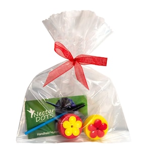 May include: A clear plastic bag with a red ribbon tied around it. Inside the bag are two yellow and red flower-shaped hummingbird feeders, a blue feeding tube, and a card with the text "Nectar Dots Handheld Hummingbird Feeder".
