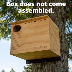 May include: A wooden birdhouse with a round entrance hole and ventilation slots, mounted on a tree. The text "Box does not come assembled" is visible at the top. The birdhouse has the text "JC's wildlife" and "Clean Oil" on the side.