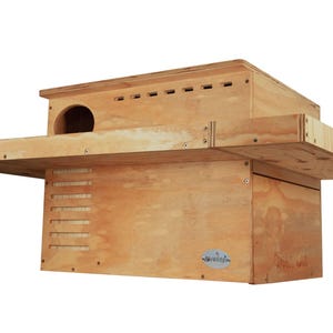 Könnte beinhalten: Ein Vogelhaus aus Holz mit natürlicher Oberfläche. Das Vogelhaus hat einen rechteckigen Körper mit einem kreisförmigen Eingangsloch und einem flachen Dach. Eine kleine Sitzstange erstreckt sich von der Vorderseite. Das Vogelhaus dient als Unterschlupf für Vögel.