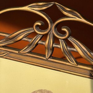 May include: A rectangular picture frame with a metallic bronze finish featuring an ornate top border decorated with raised leaf and scroll motifs. The frame holds a light-coloured insert displaying a single flower illustration in the centre.