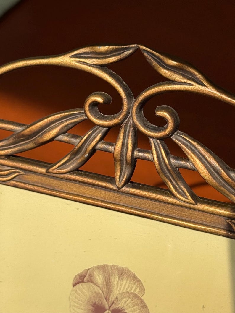 May include: A rectangular picture frame with a metallic bronze finish featuring an ornate top border decorated with raised leaf and scroll motifs. The frame holds a light-coloured insert displaying a single flower illustration in the centre.