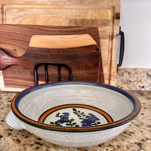 May include: A speckled ceramic bowl with a blue, orange, and green rim. The bowl features a floral design in the center. A wooden cutting board and a black metal rack are in the background.
