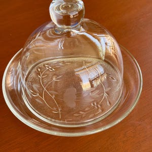 May include: A clear glass butter dish with a dome-shaped lid featuring a frosted floral etching. The lid has a round knob handle on top, and the base is a circular plate with a smooth rim. The glass is transparent with etched botanical details.