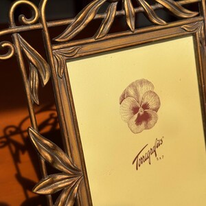 May include: A bronze-toned metal picture frame with an ornate leaf and scrollwork border. The frame holds a light yellow insert featuring a brown pansy illustration and the text Terragraphics 5x7, indicating a 12.7 cm by 17.8 cm photo size.