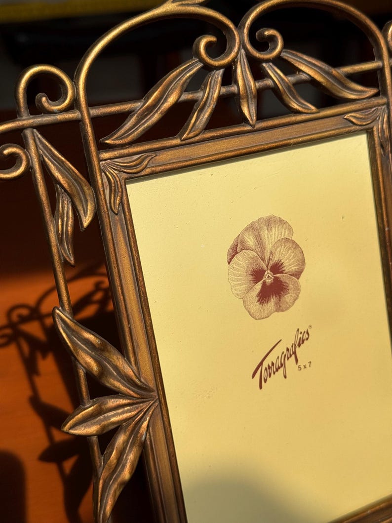 May include: A bronze-toned metal picture frame with an ornate leaf and scrollwork border. The frame holds a light yellow insert featuring a brown pansy illustration and the text Terragraphics 5x7, indicating a 12.7 cm by 17.8 cm photo size.