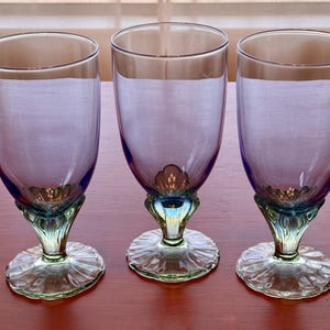 Puede incluir: Tres copas a juego con cuencos de color morado translúcido y bases de color verde claro. Cada copa presenta un diseño festoneado en forma de concha en la base del cuenco y un pie estriado en forma de pétalo, creando un conjunto coherente.