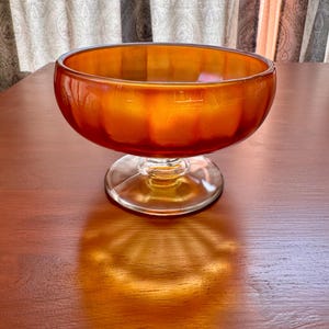 Op de afbeelding: Een amberkleurige glazen kom met een afgeronde vorm, geplaatst op een heldere, cirkelvormige basis. Het oppervlak van de kom reflecteert licht, waardoor een warme gloed ontstaat. De steel is kort en de kom staat op een houten oppervlak.