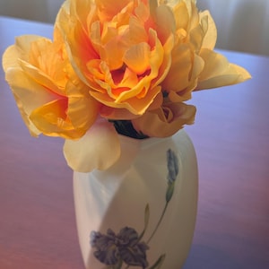 May include: A white ceramic vase with a subtle twisted geometric shape and a painted purple iris flower design, containing a cluster of vibrant yellow and orange artificial peony flowers with layered, ruffled petals and soft edges.