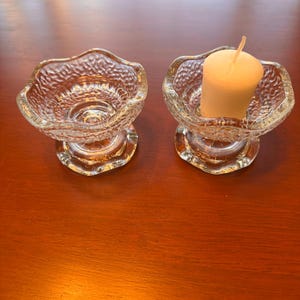 May include: Two clear glass candle holders with a textured, pebbled surface and a scalloped rim. One holder is empty, while the other contains a small, cylindrical white pillar candle with a visible wick centered in the glass bowl.