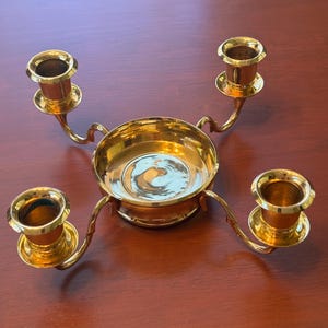 May include: A polished gold-tone metal candelabra featuring a central circular bowl base with four curved arms extending outwards, each topped with a cylindrical candle holder cup designed to hold a single taper candle in each position.