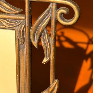 May include: A rectangular picture frame with a metallic gold finish and a distressed texture. The frame features decorative raised leaf motifs along the vertical edges and a curved scroll design at the top corner, surrounding a plain cream insert.