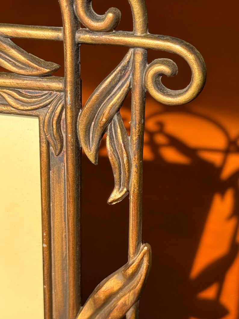 May include: A rectangular picture frame with a metallic gold finish and a distressed texture. The frame features decorative raised leaf motifs along the vertical edges and a curved scroll design at the top corner, surrounding a plain cream insert.
