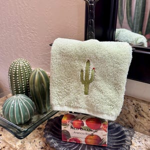 May include: A light green hand towel with an embroidered cactus design hanging on a dark metal stand. Below the towel sits a rectangular bar of Red Orange Arancio Rosso vegetable soap in a decorative dark metal dish with a scalloped edge.