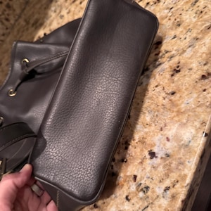 May include: A dark brown handbag with a textured surface and a rectangular base. The bag features gold-toned metal hardware, including circular grommets for the straps, and a structured design with visible stitching along the bottom edges.