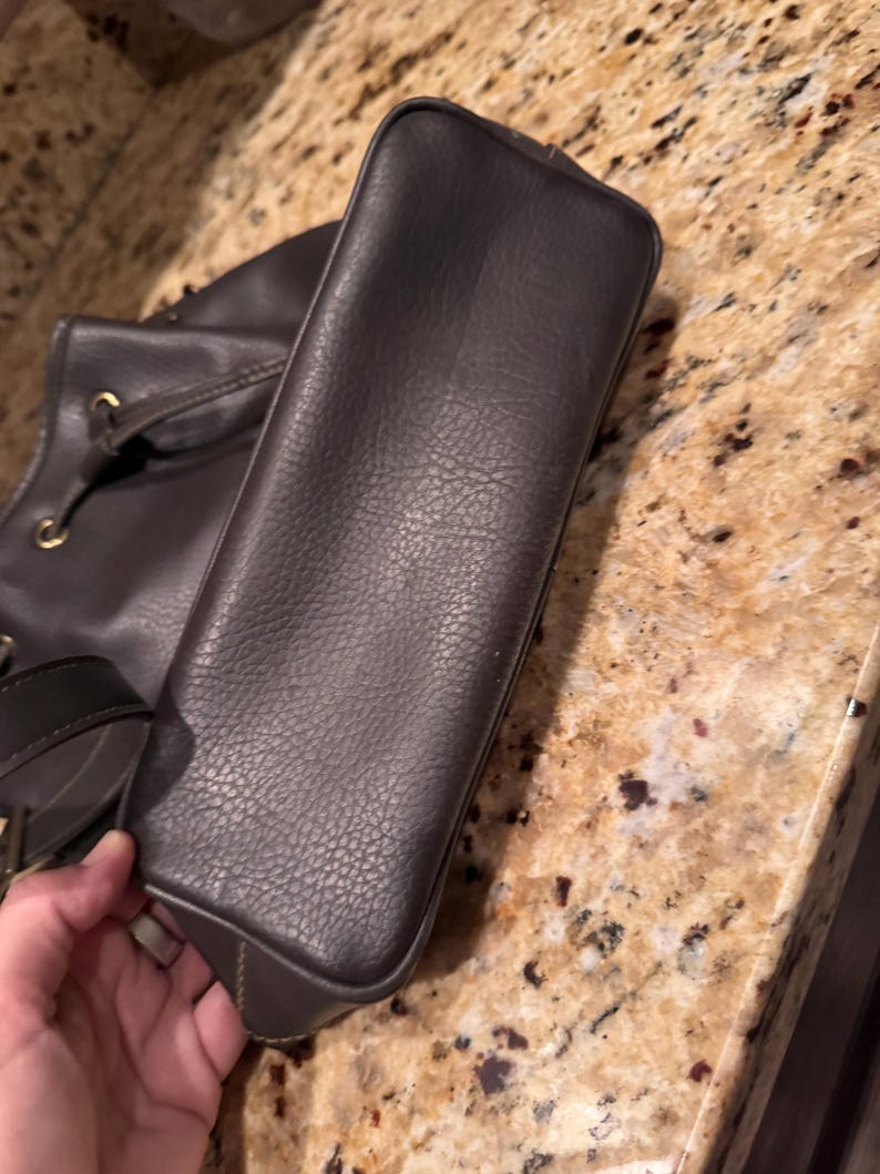 May include: A dark brown handbag with a textured surface and a rectangular base. The bag features gold-toned metal hardware, including circular grommets for the straps, and a structured design with visible stitching along the bottom edges.