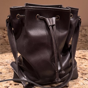 May include: A dark grey bucket bag with a textured surface, featuring a drawstring closure with matching leather cords, brass-toned metal eyelets, and an adjustable shoulder strap with a metal buckle for carrying.