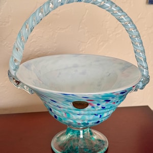 Op de afbeelding: Een decoratieve glazen mand met een handvat. De mand heeft een witte binnenkant en een turquoise buitenkant met blauwe, roze en groene spikkels. Het handvat is van gedraaid lichtblauw glas. De mand staat op een heldere, groen getinte voet.