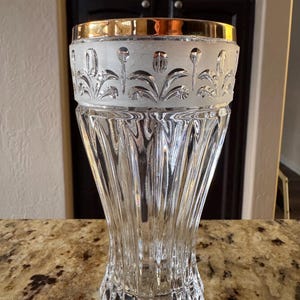 May include: A clear glass vase with a gold rim and frosted floral design. The vase has a fluted body and a decorative base. The top of the vase has a gold band. The vase is sitting on a brown and tan surface.