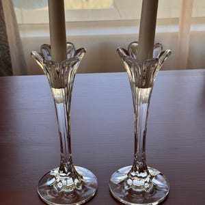 May include: A pair of clear glass candle holders with a fluted, petal-like top design and a slender, vertical ribbed stem. Each holder features a circular base and contains a single, tall, slender, off-white taper candle inserted inside.
