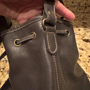 May include: Dark brown leather drawstring bag with brass-coloured hardware. The bag has a textured surface and visible stitching. The handle is attached with a brass-coloured ring.