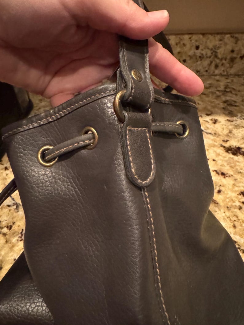 May include: Dark brown leather drawstring bag with brass-coloured hardware. The bag has a textured surface and visible stitching. The handle is attached with a brass-coloured ring.