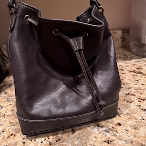 May include: A dark brown leather bucket bag with a drawstring closure and a shoulder strap. The bag features the brand name ESPRIT embossed on the lower front panel. It has brass-toned metal hardware, including eyelets for the drawstring cord.