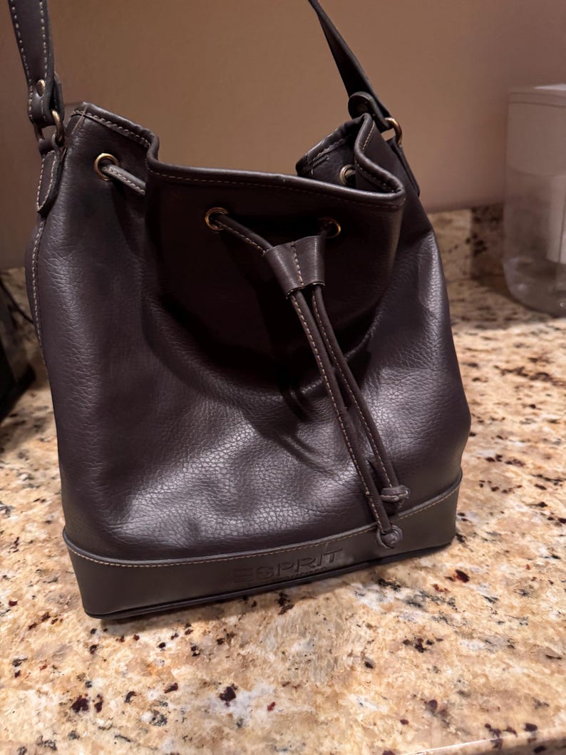 May include: A dark brown leather bucket bag with a drawstring closure and a shoulder strap. The bag features the brand name ESPRIT embossed on the lower front panel. It has brass-toned metal hardware, including eyelets for the drawstring cord.