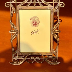 May include: A rectangular metal picture frame with an antique bronze finish and ornate leaf-shaped accents. The frame features a central insert with a printed pansy illustration and the text Terragrafics 5x7, indicating a 12.7 by 17.8 cm size.