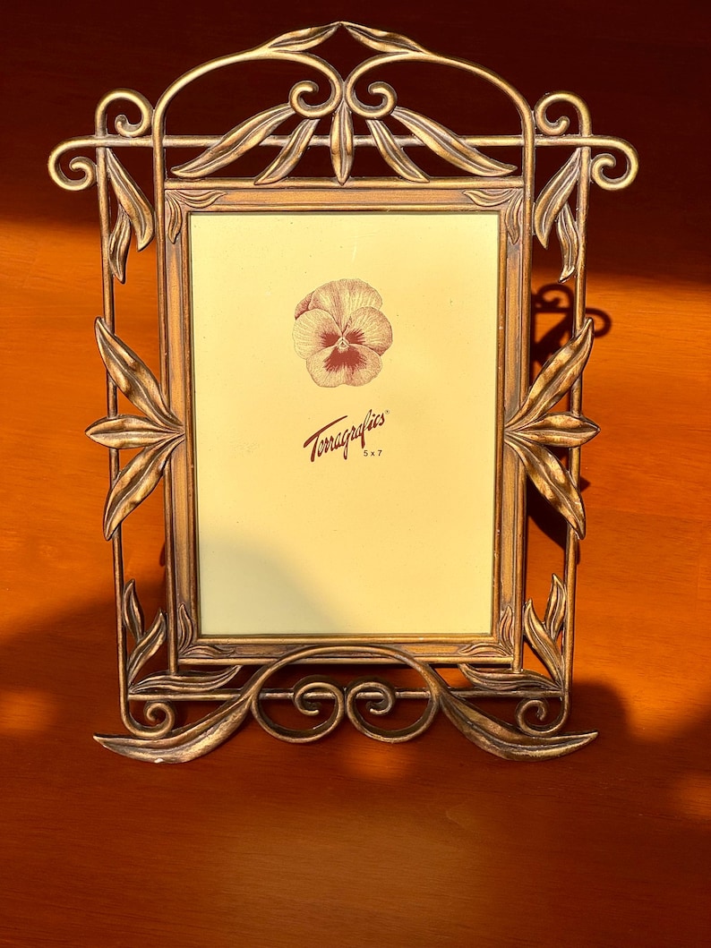 May include: A rectangular metal picture frame with an antique bronze finish and ornate leaf-shaped accents. The frame features a central insert with a printed pansy illustration and the text Terragrafics 5x7, indicating a 12.7 by 17.8 cm size.