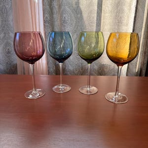 May include: A set of four stemmed wine glasses featuring rounded bowls in distinct translucent colors: deep plum, slate blue, olive green, and amber. Each glass has a clear, slender stem and a circular base, standing upright.